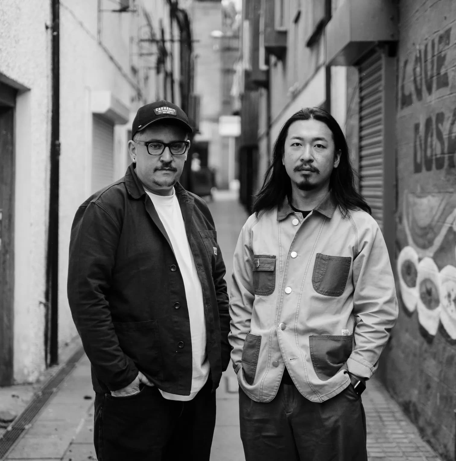 Two men stand together in a narrow urban alley, looking directly at the camera with serious expressions.