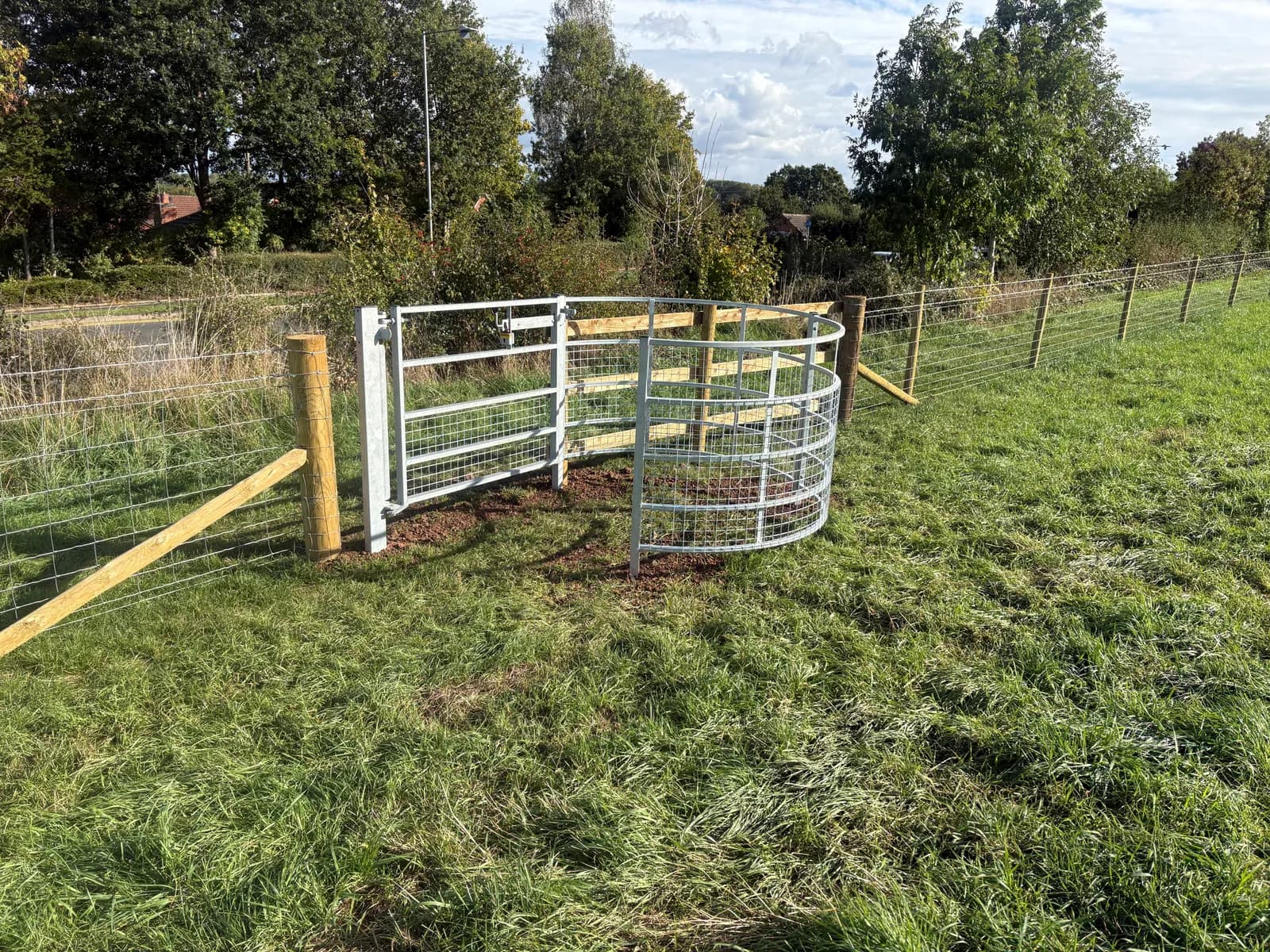 A new metal gate and curved enclosure are installed in a grassy field with a wire fence stretching into the distance.