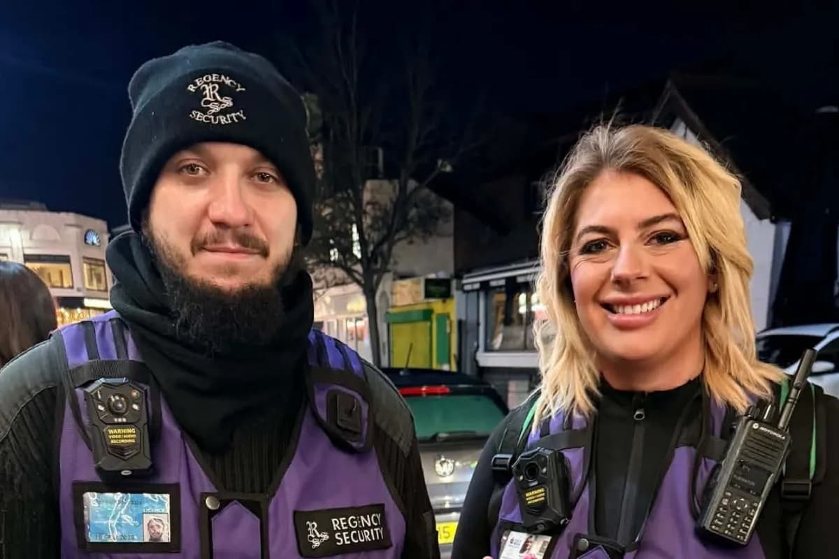 A man and a woman in purple security vests and black attire smile at the camera at night.