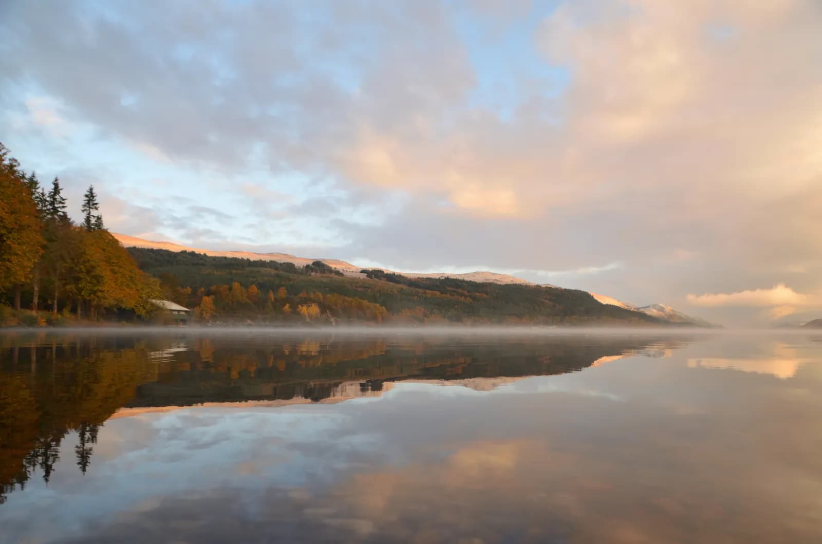 A scenic view of a calm lake with mist rising from its surface, surrounded by hills covered in autumn trees and reflected clouds at sunrise.