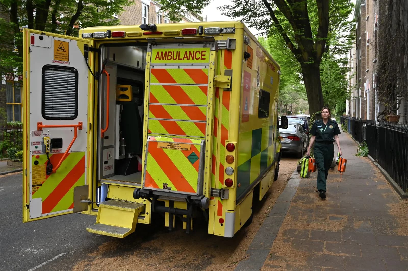 A female paramedic in a dark green uniform walks along a sidewalk carrying two medical bags, passing a yellow ambulance with its rear doors open.
