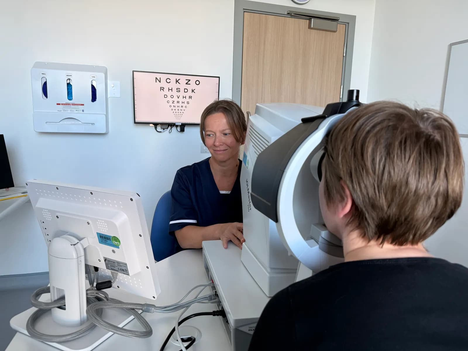 A female optometrist wearing blue scrubs looks on as a patient seated with their back to the camera has their eyes examined by a large white medical device.