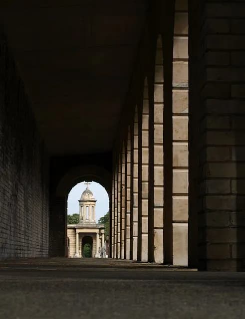 A long, dark arched corridor with a distant stone building with a dome and cupola visible through the archway at the end.
