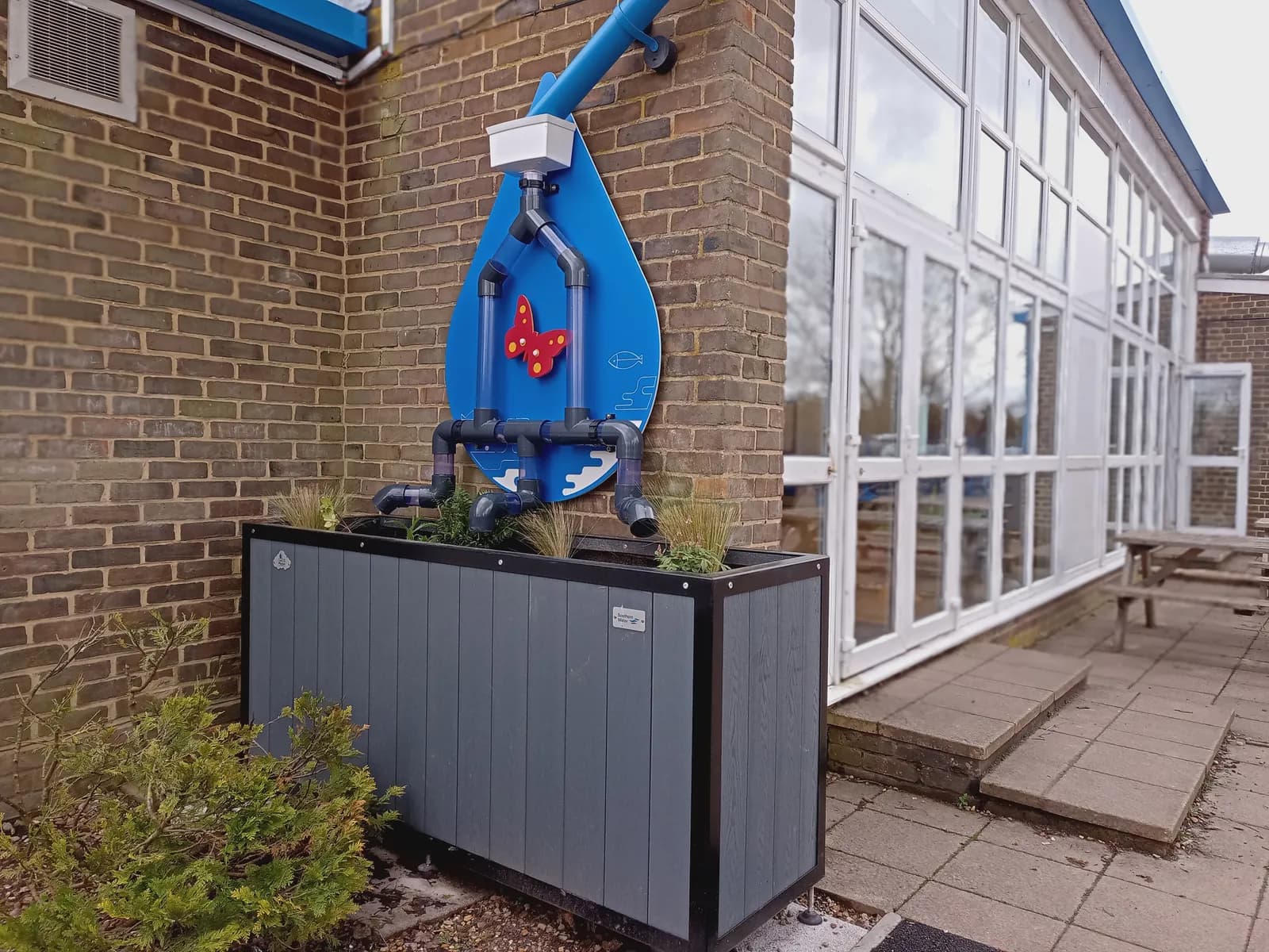 A large blue rain gauge in the shape of a water droplet, with pipes leading down to a planter box, is affixed to a brick wall next to a building with many windows.