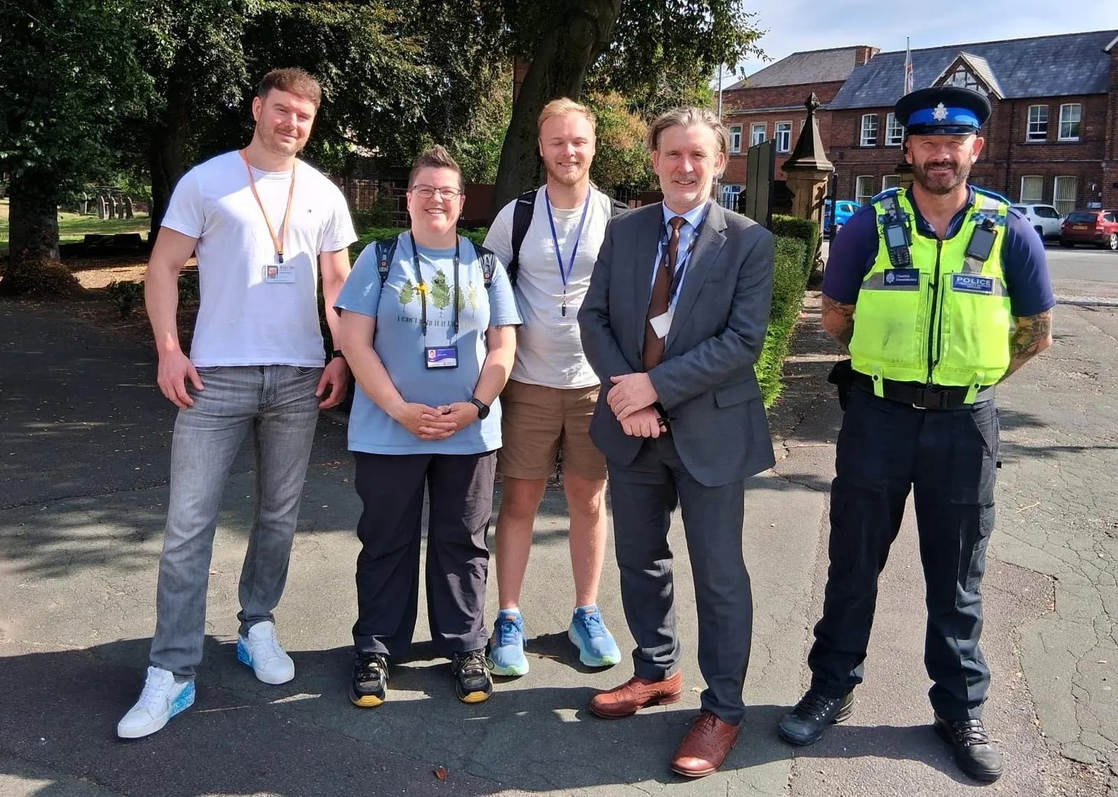 Five adults, three men, one woman, and a police officer, stand smiling in a row on a sunny day with trees and buildings in the background.