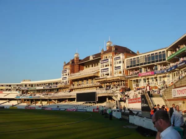 A wide shot of a cricket stadium with a large, ornate grandstand filled with spectators under a clear blue sky.