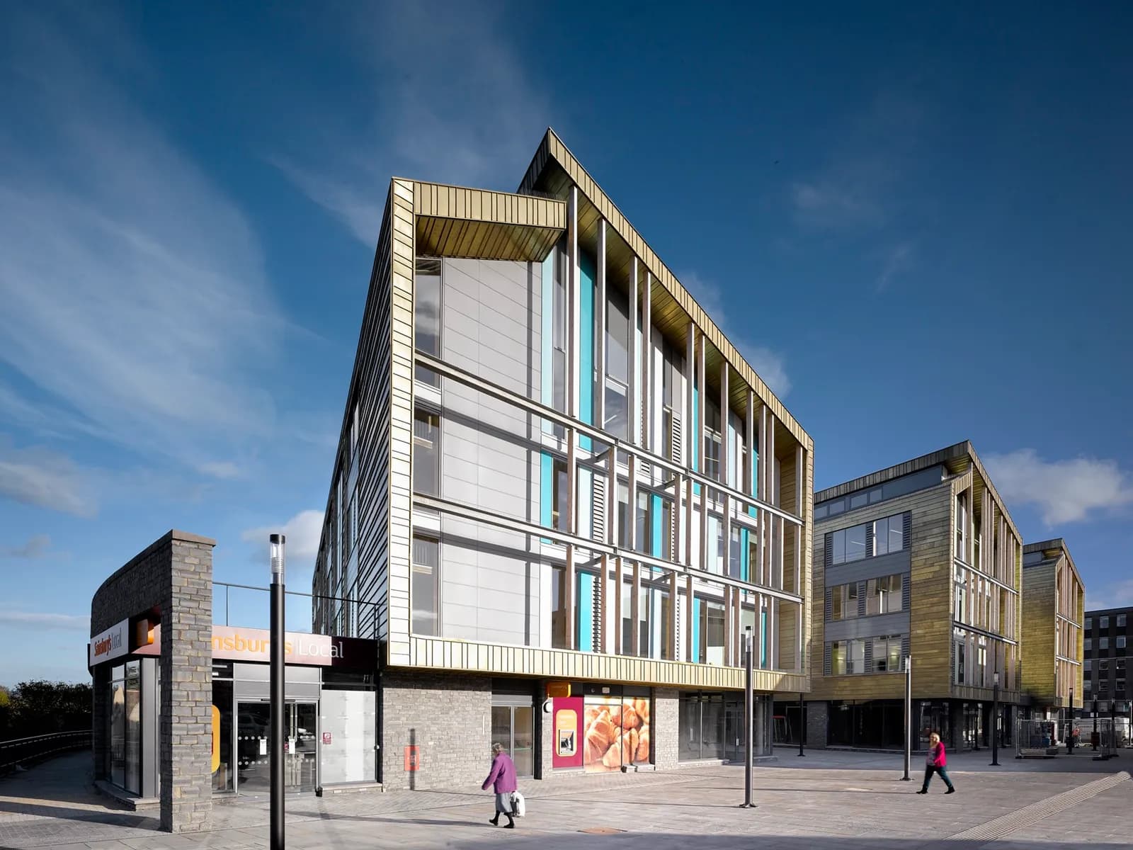 A modern building with a gold-colored sloped roof and facade, featuring large windows and some blue accents, stands next to a smaller, rounder brick structure with a "Sainsbury's Local" sign, with people walking on the paved area in front.