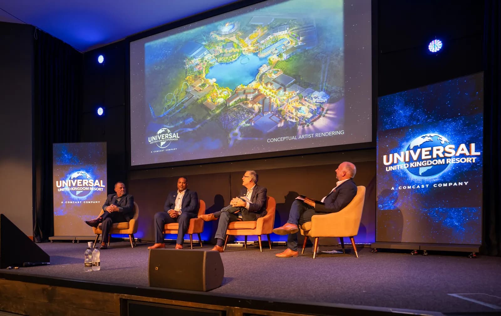Four men sit on orange chairs on a stage in front of a large screen displaying a conceptual art rendering of a Universal resort.