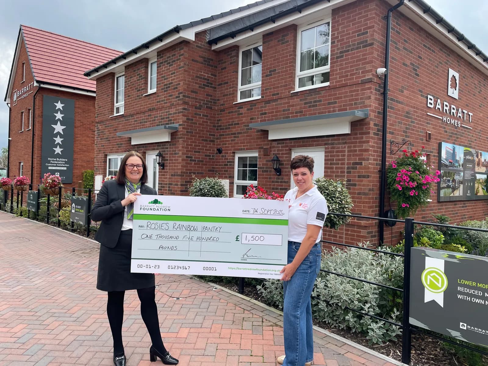 Two women, one in a business suit and the other in jeans and a white polo, stand in front of new brick houses, holding a large check for £1,500 made out to Rosie's Rainbow Pantry.