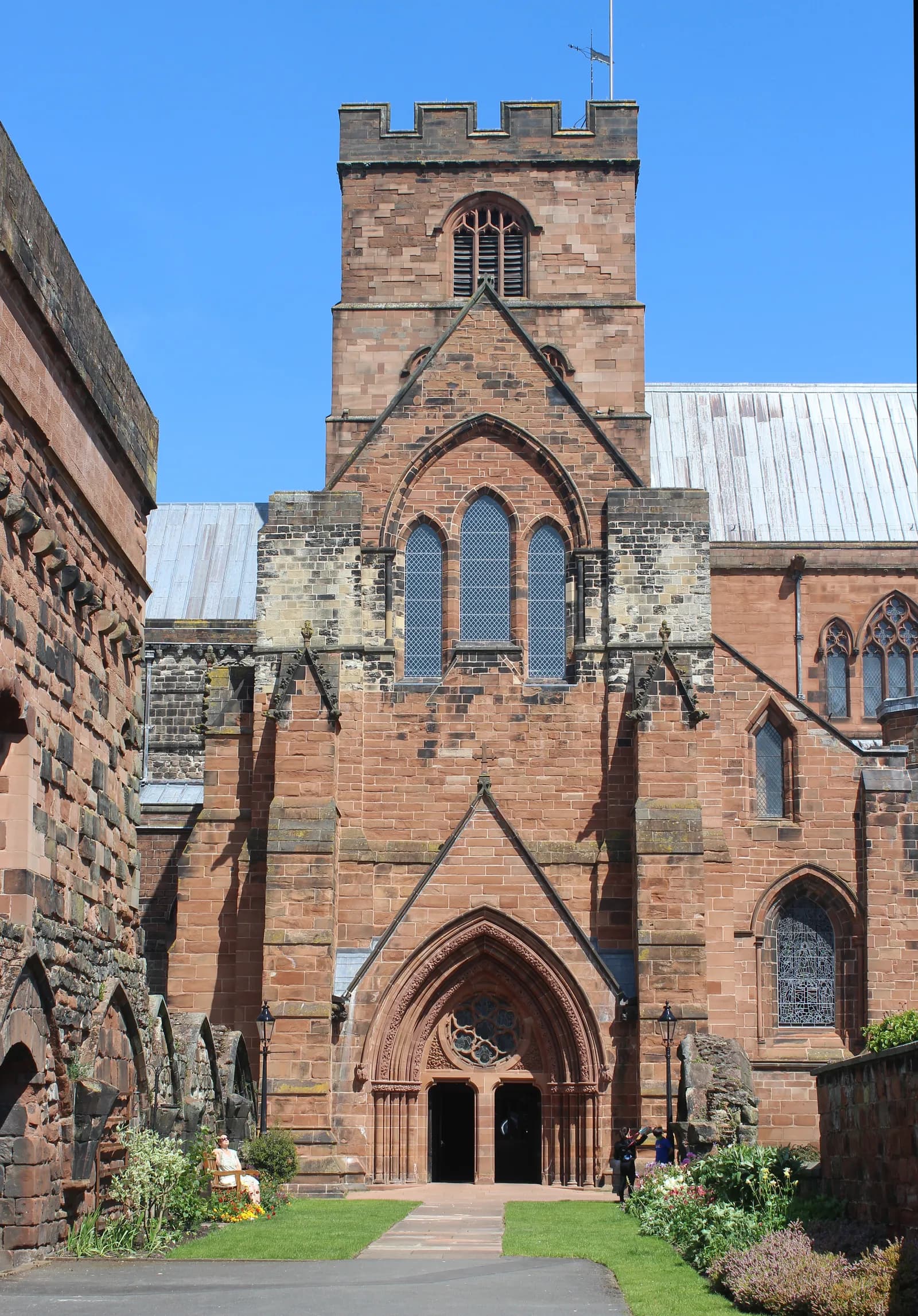 A towering Cathedral with intricate red brickwork, arched windows, and a prominent entrance, set against a clear blue sky, with some greenery and people near the base.