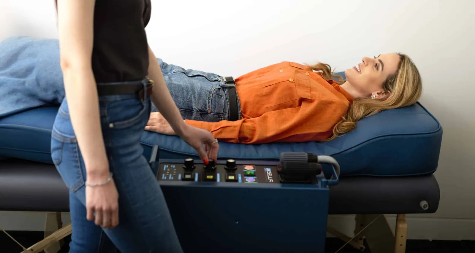 A woman in a black shirt and jeans operates a medical device next to another woman lying on a blue padded bed.