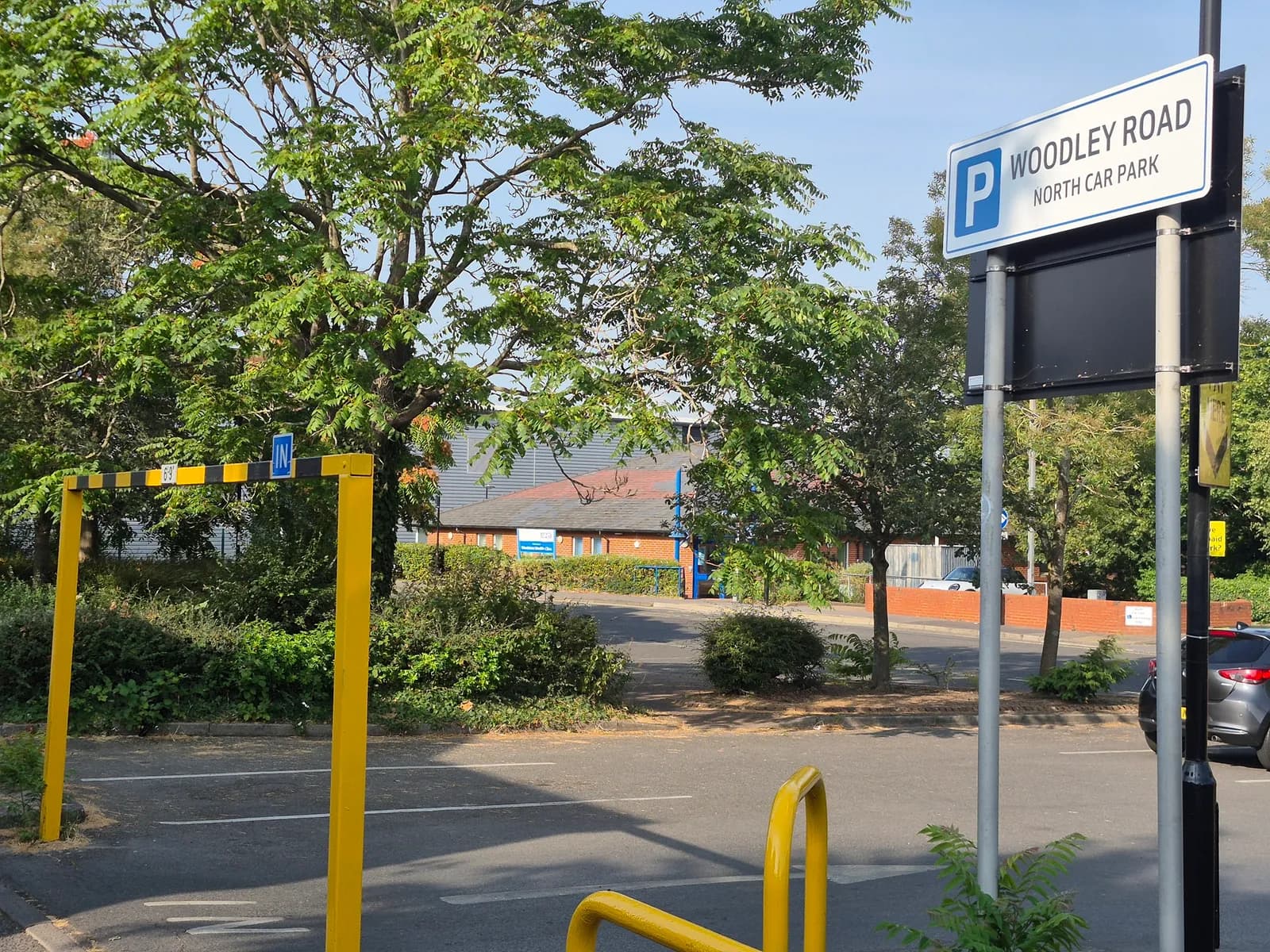 A car park entrance with a yellow height restriction bar, an 'IN' sign, and a 'WOODLEY ROAD NORTH CAR PARK' sign on a pole.