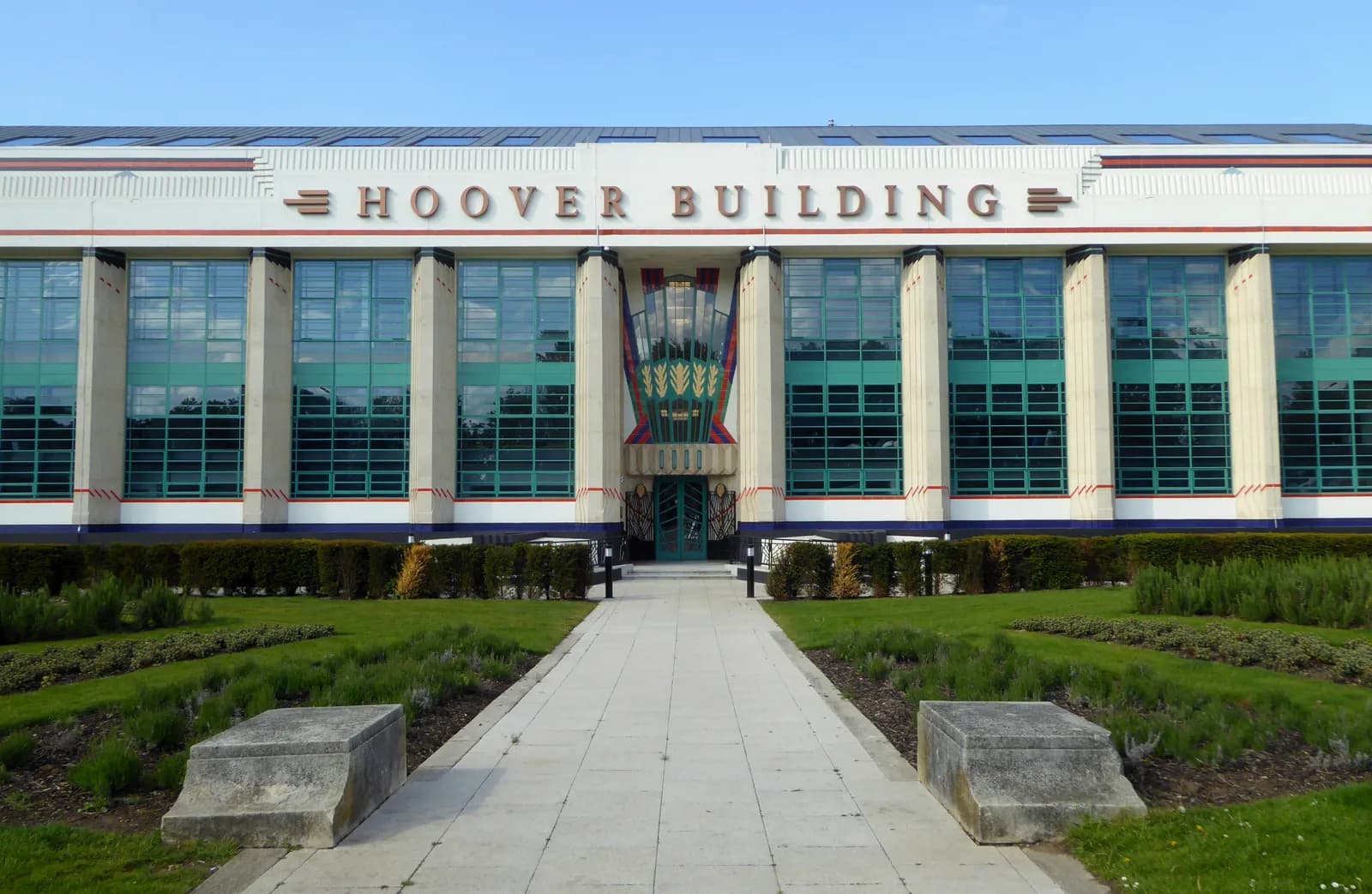 The Hoover Building, a white Art Deco building with prominent green-framed windows, stands under a clear blue sky, with a paved walkway leading through manicured green lawns toward its central entrance.