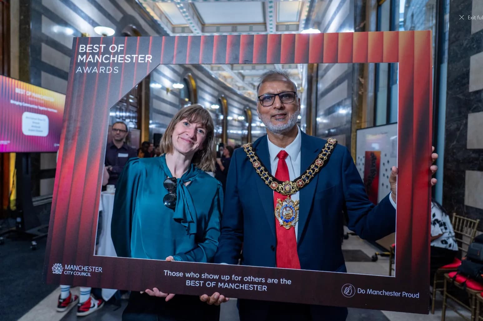 A woman and a man in a mayoral chain stand behind a large brown frame with text promoting the Best of Manchester Awards.