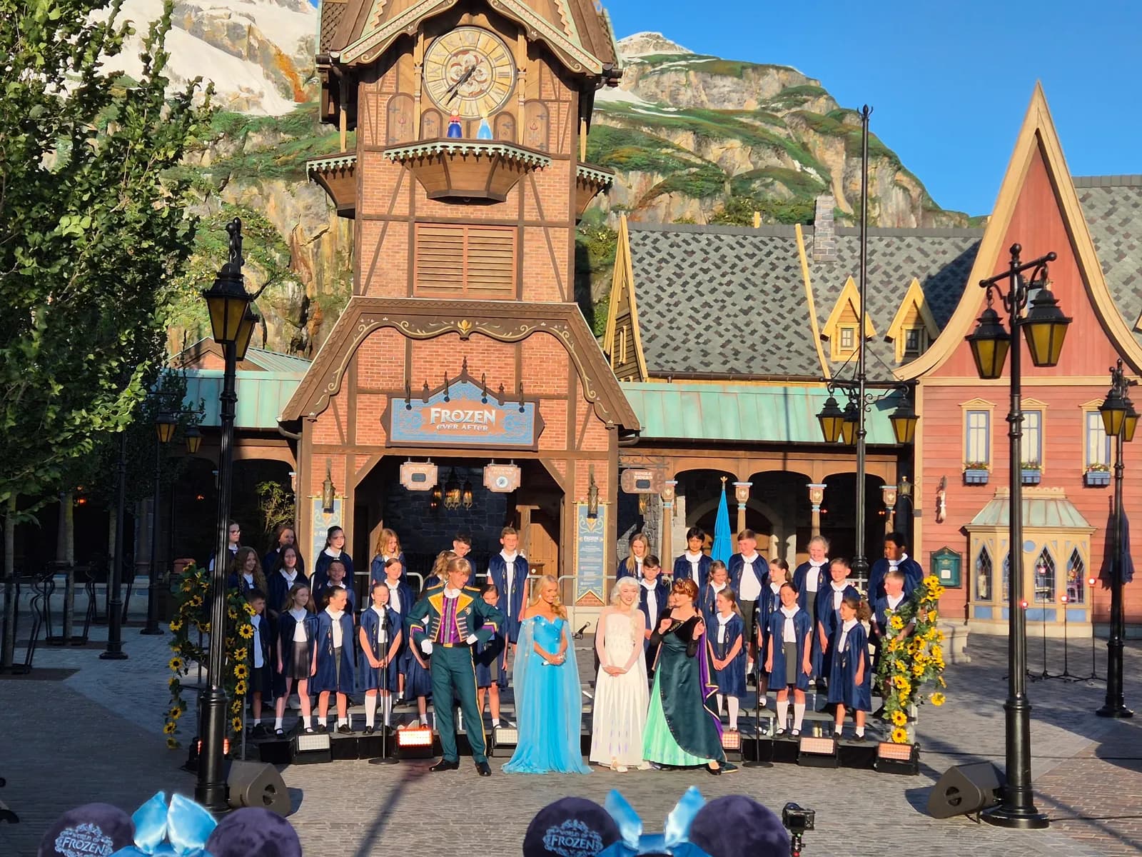 A live performance with actors dressed as characters from Disney's Frozen, accompanied by a children's choir, takes place in front of a themed building with a clock tower.