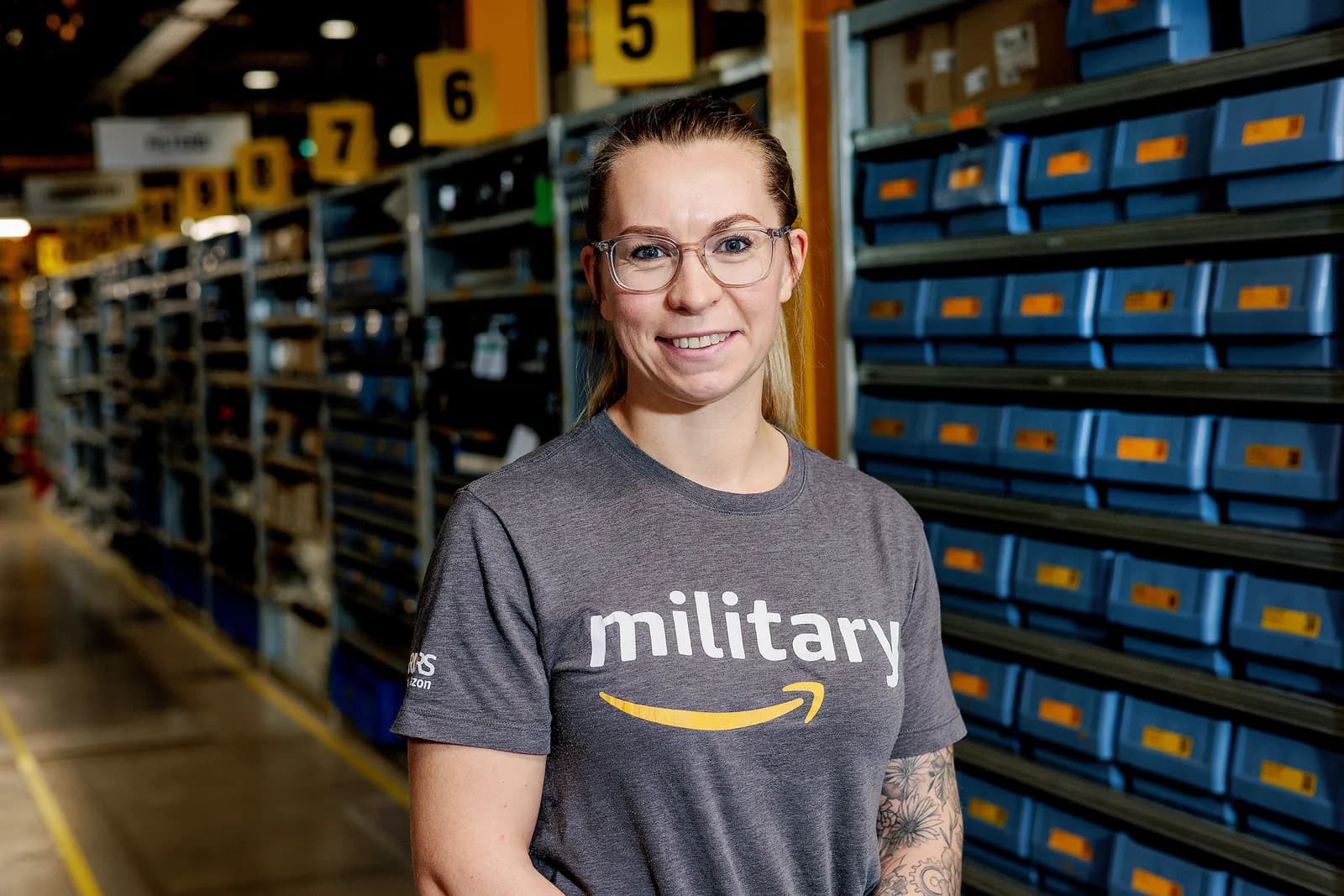 A woman wearing glasses and an Amazon military shirt smiles at the camera in a warehouse setting with shelves of blue bins behind her.