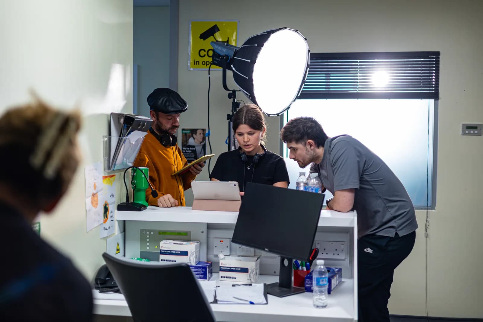 Three people, two men and one woman, stand behind a white counter with equipment like a tablet, monitor, and light reflector, appearing to be on a film set.