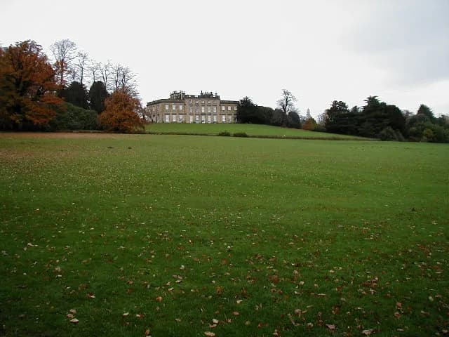 A grand, multi-story stone manor house sits atop a grassy hill, surrounded by trees with some autumn foliage, under a cloudy sky.
