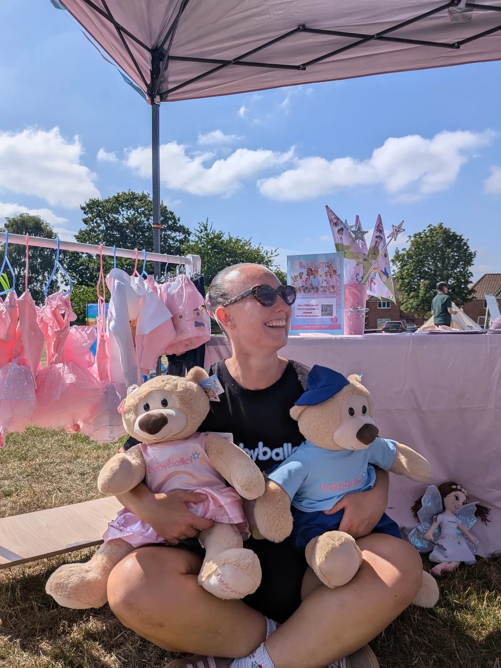 A smiling woman wearing sunglasses and a black t-shirt is sitting on the ground, holding two large teddy bears in her lap, with a pink market stall and green trees in the background.
