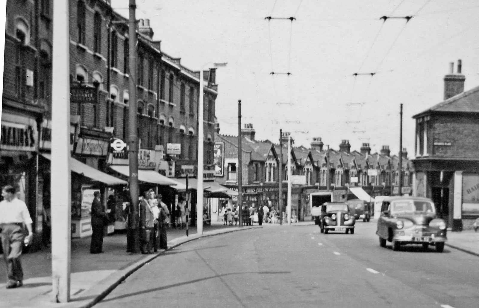 A black and white photograph shows a street scene in a bustling town with people walking on the sidewalks and several cars driving on the road.
