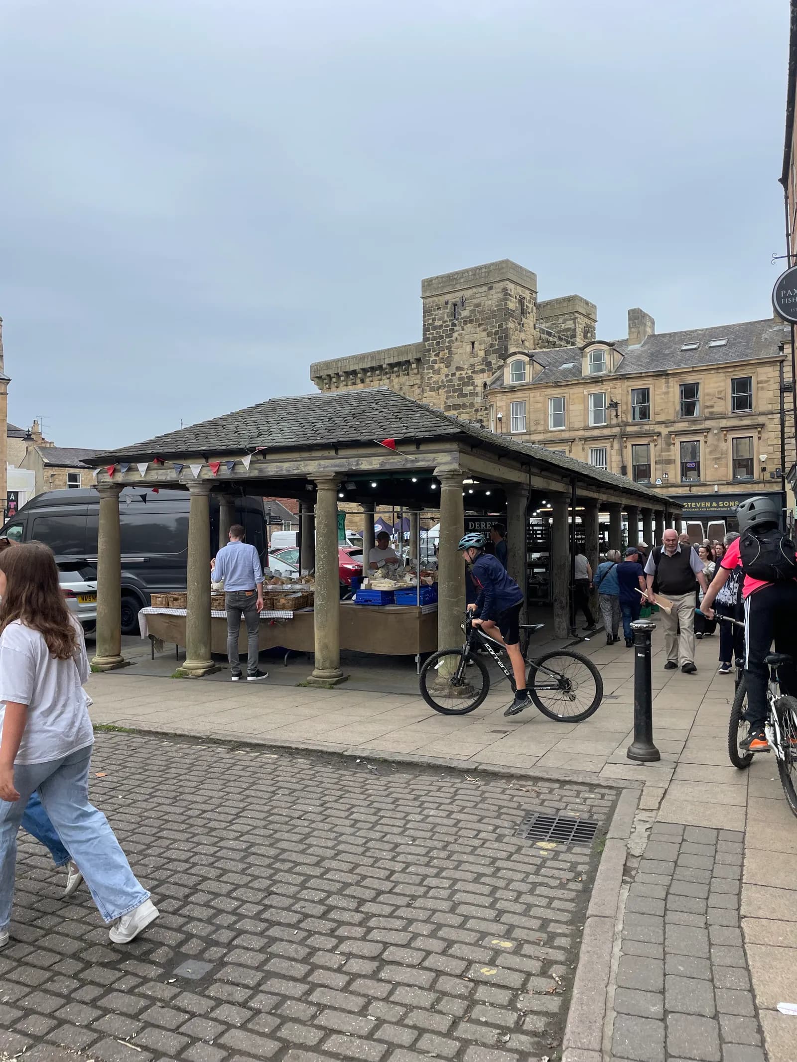 A boy on a bicycle riding past an open-air market with stone columns and a tiled roof, with historic buildings and people in the background.