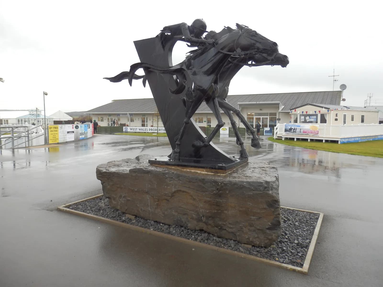 A large black metal sculpture of a horse and jockey racing, set on a stone base, with buildings and a wet paved area in the background under an overcast sky.