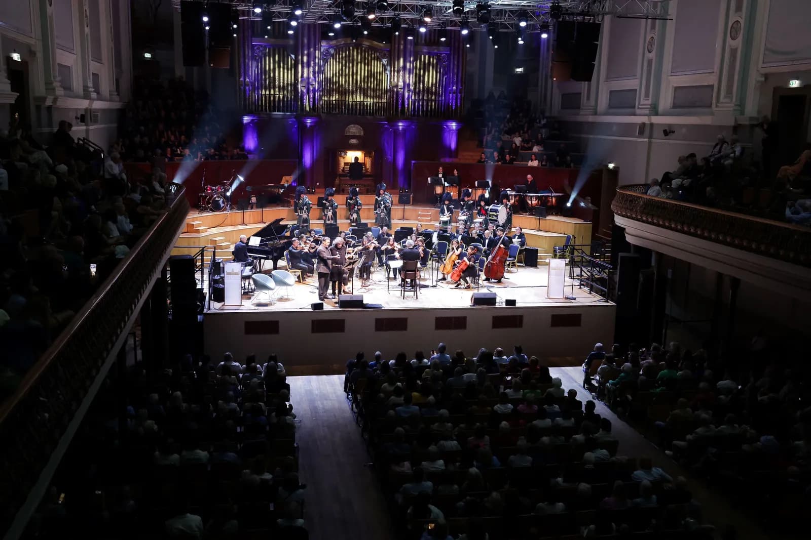 An overhead view of a large orchestra performing on a brightly lit stage in a grand concert hall with a large pipe organ visible in the background.