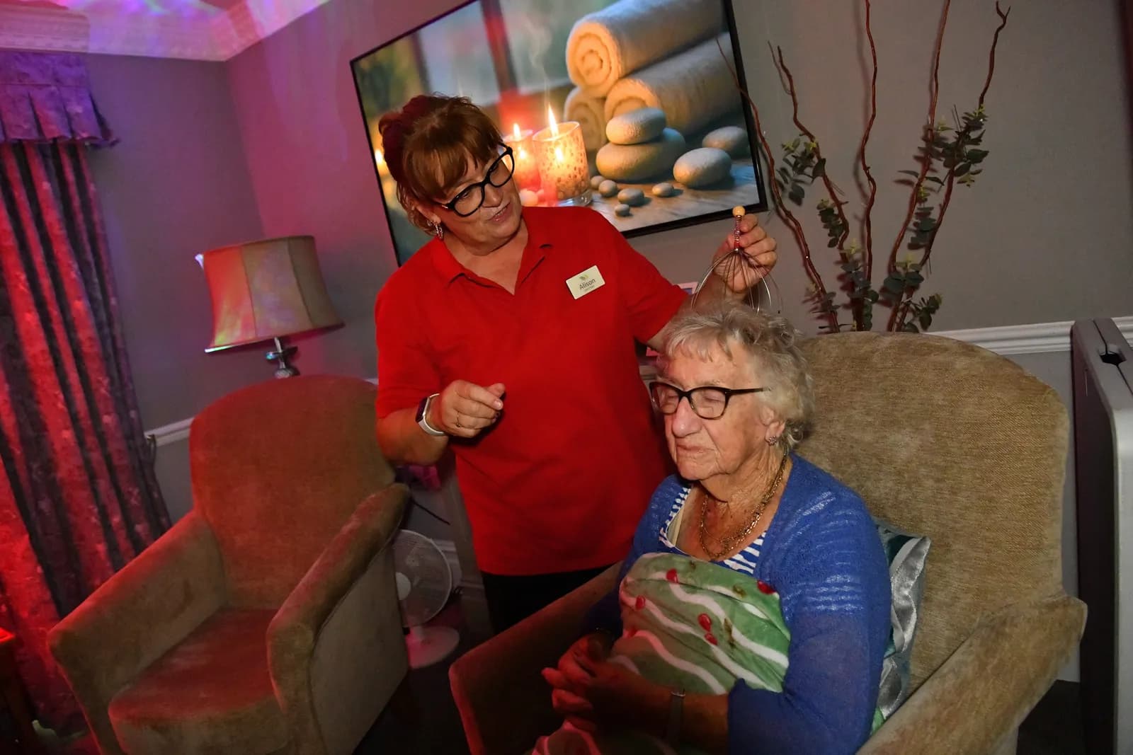 A woman in a red shirt gives a scalp massage to an older woman wearing glasses, who is sitting in an armchair with her eyes closed.