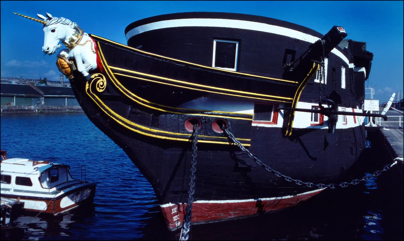 A unicorn figurehead adorns the bow of a dark-hulled ship with gold trim, docked beside a smaller white boat in blue water.