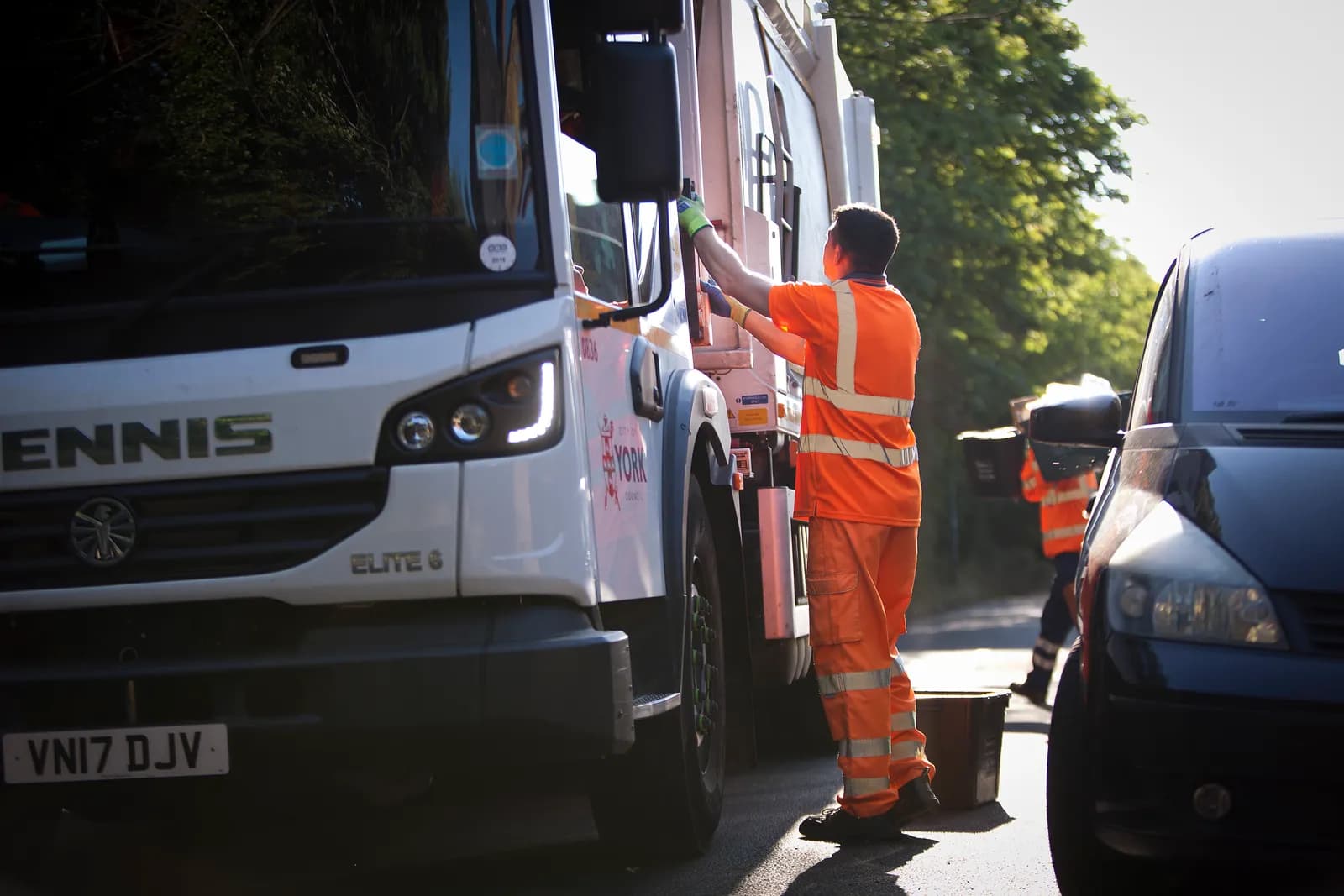 A person in an orange high-visibility uniform reaching up to the side of a white garbage truck, with a dark car partially visible on the right.