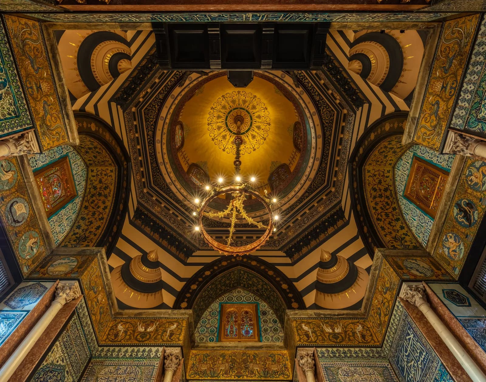 A grand, intricately decorated ceiling and dome featuring rich gold, navy, and teal patterns, with a large ornate chandelier hanging from the center.