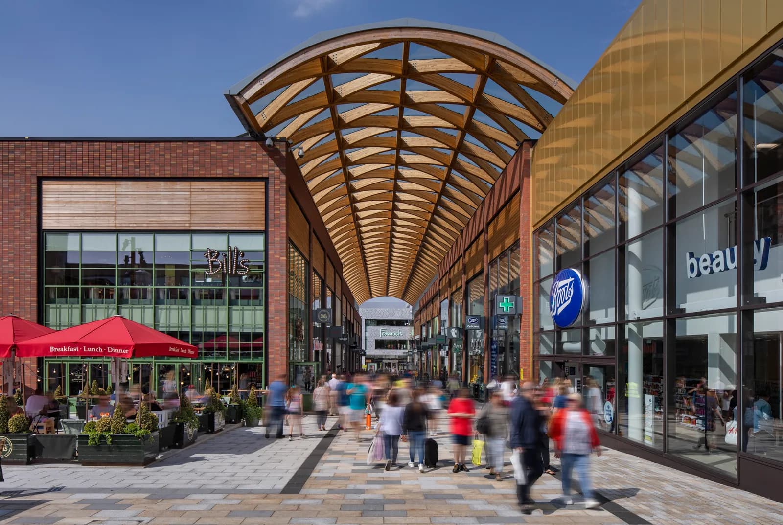 A bustling outdoor shopping street featuring retail stores, restaurants with outdoor seating under red umbrellas, and a prominent arched wooden canopy overhead.