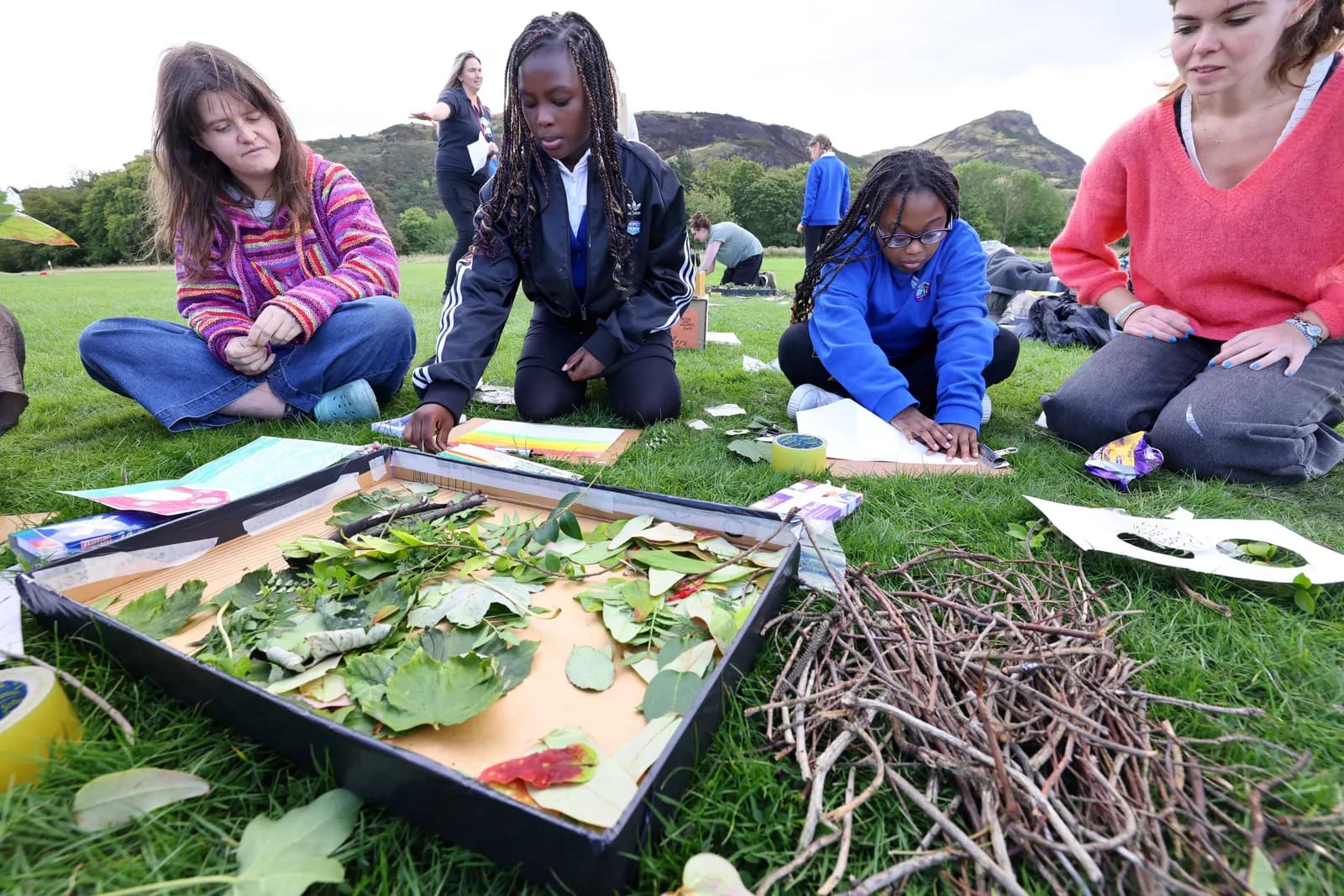 Several people are sitting on grass, engaged in an outdoor craft activity, surrounded by natural materials like leaves and twigs.