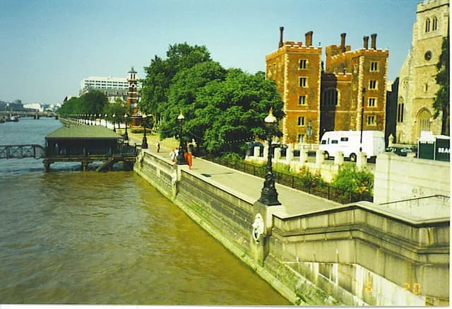A long shot of the Thames River embankment, showing brick buildings and churches on the shore, with tree-lined pedestrian areas and a small boat shed by the water.