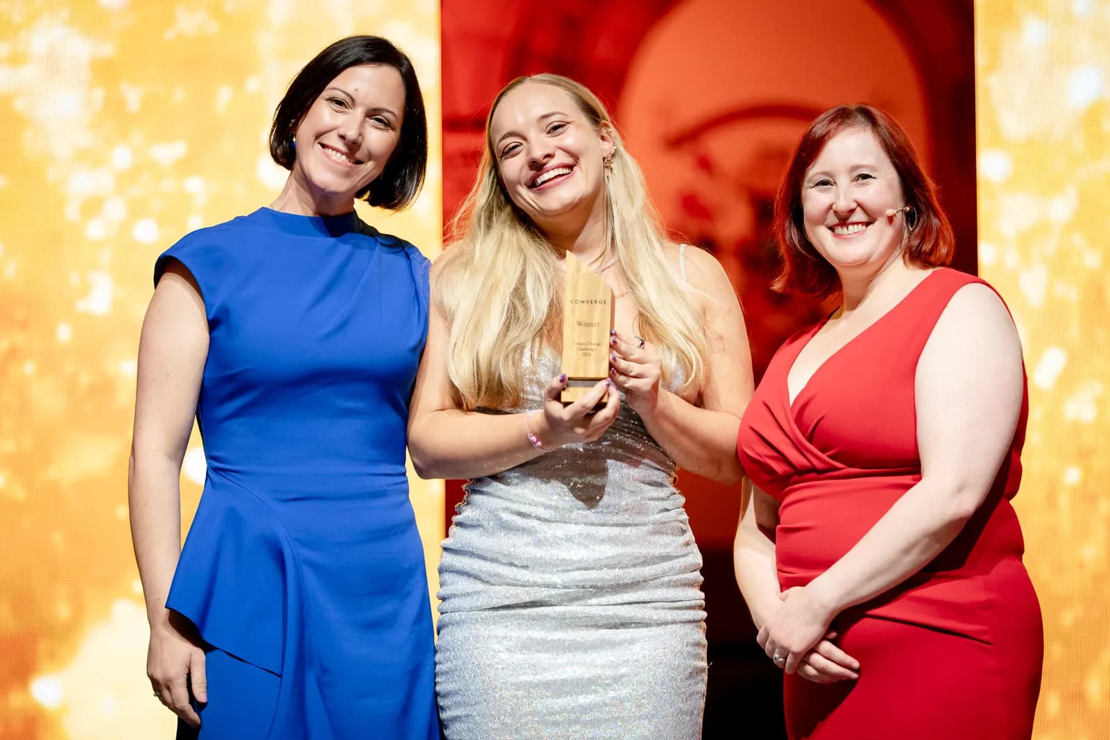 Three smiling women stand on a stage with a brightly lit background, the woman in the center holding a wooden award.