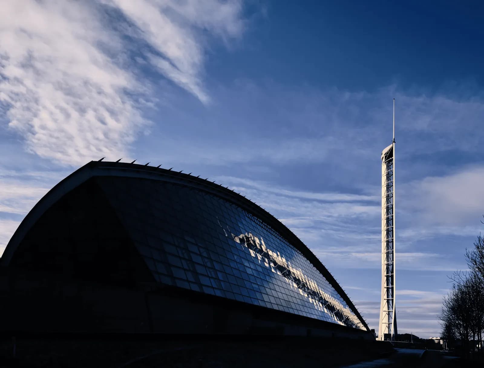 A long, curvilinear building with a dark facade and reflective windows sits next to a tall, slender tower against a bright blue sky with wispy clouds.