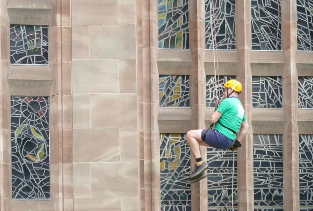 A person in a yellow helmet and green shirt rappels down a stone building with stained glass windows.