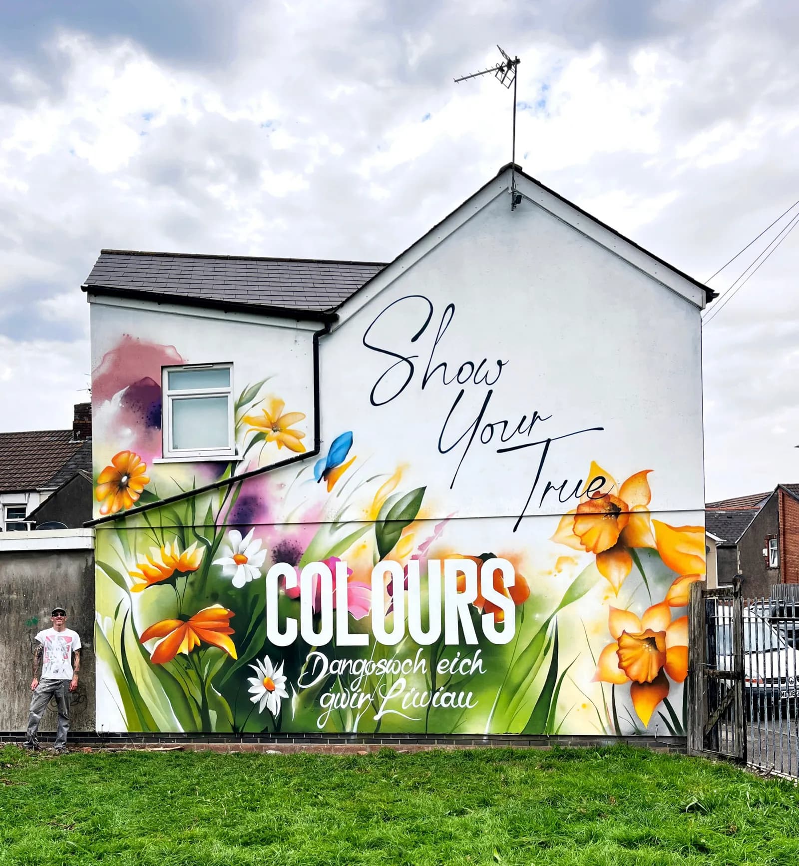 A man stands next to a building with a vibrant floral mural that reads "Show Your True COLOURS" with a Welsh translation below.