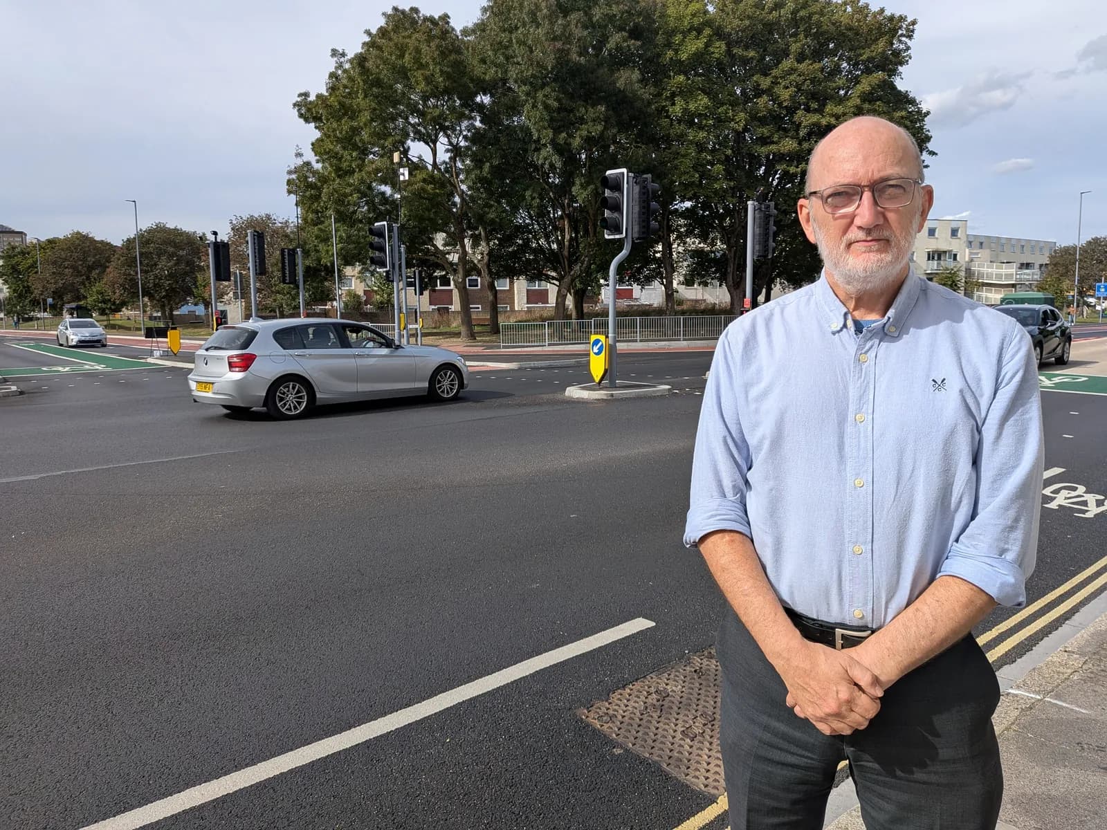 A man wearing glasses and a light blue shirt stands on the side of a road, looking at the camera, with cars and trees in the background.