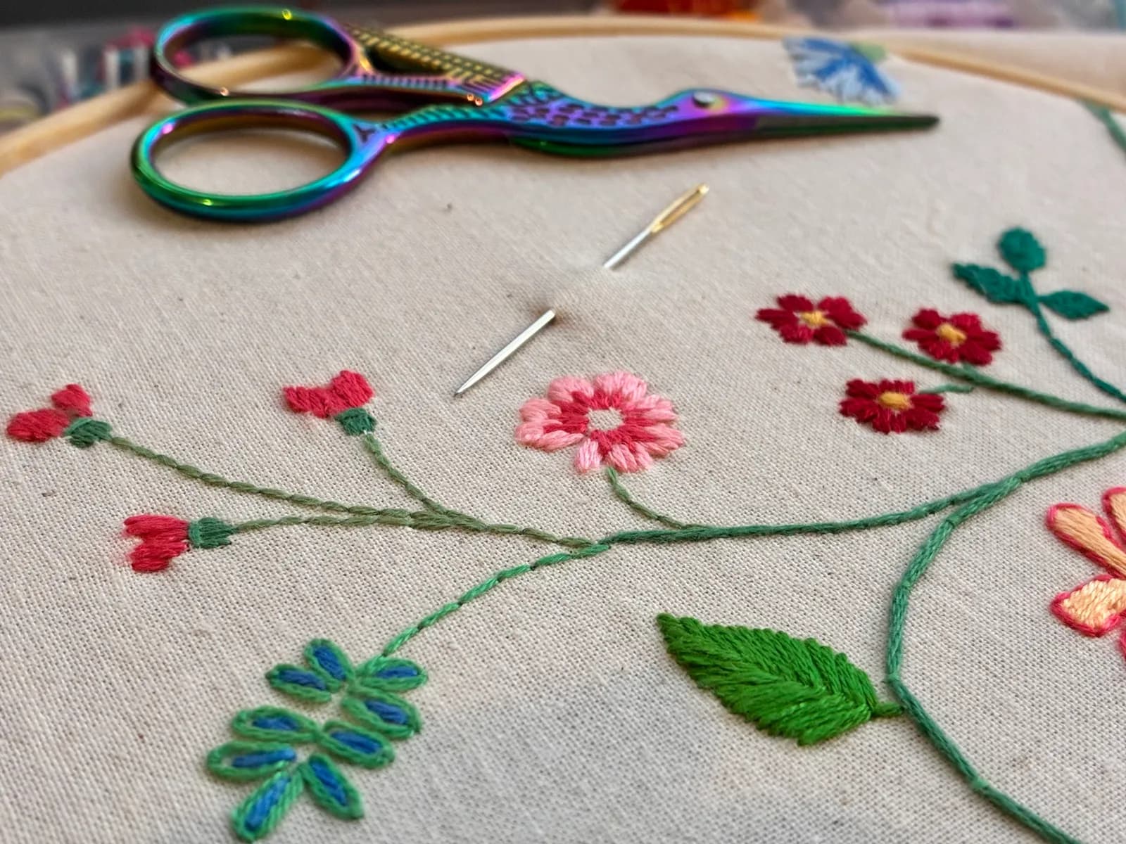 A close-up view of an embroidery project featuring colorful floral designs on a light fabric, with iridescent scissors and sewing needles resting on it.