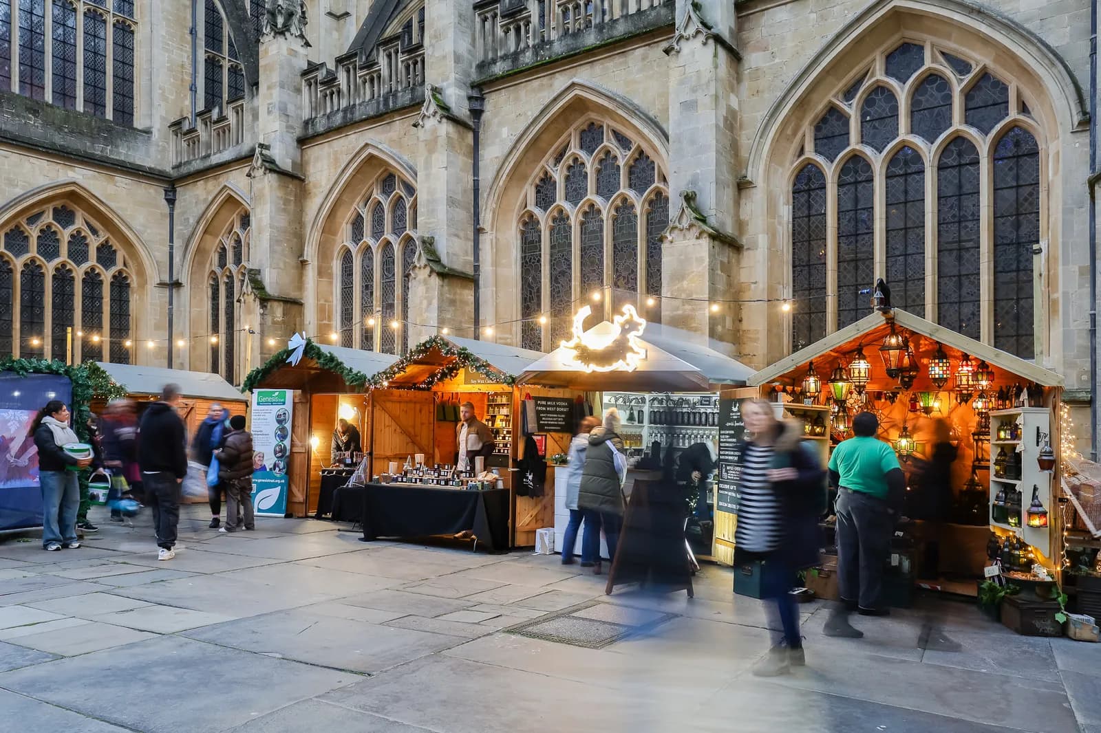 A bustling outdoor Christmas market with several wooden stalls illuminated by festive lights, set against the backdrop of an old stone church with large gothic windows.