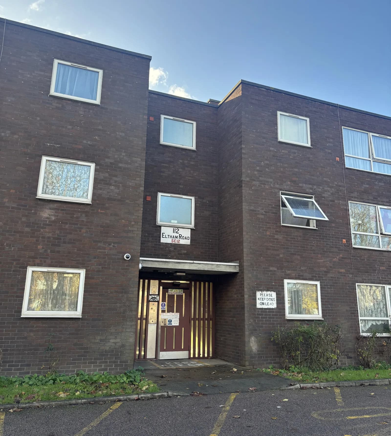 A three-story brick apartment building with a red and gold door and a sign that reads "112 Eltham Road SE12" above the entrance.