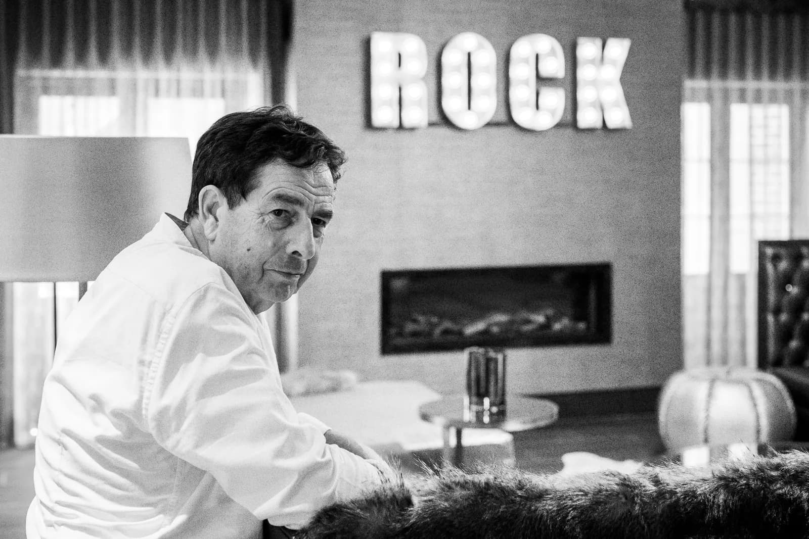 A man in a white shirt looks over his shoulder at the camera in a room with a decorative 'ROCK' sign on the wall.