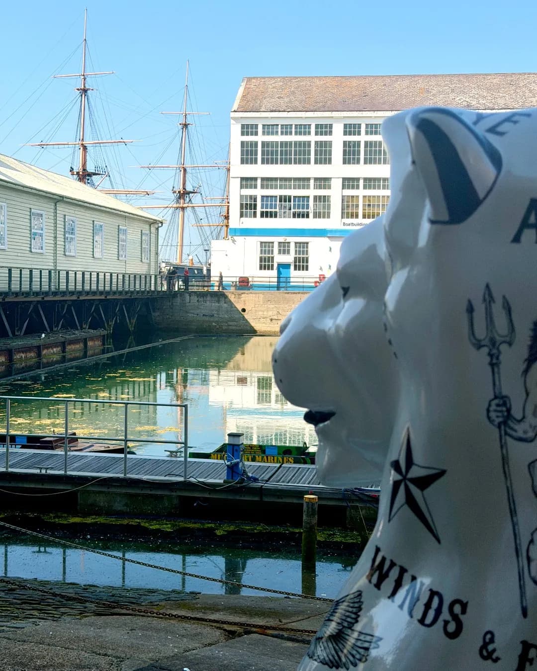A large, white pig statue with a nautical tattoo design on its side stands in the foreground, facing a bustling harbor with historic buildings and tall ships under a clear sky.