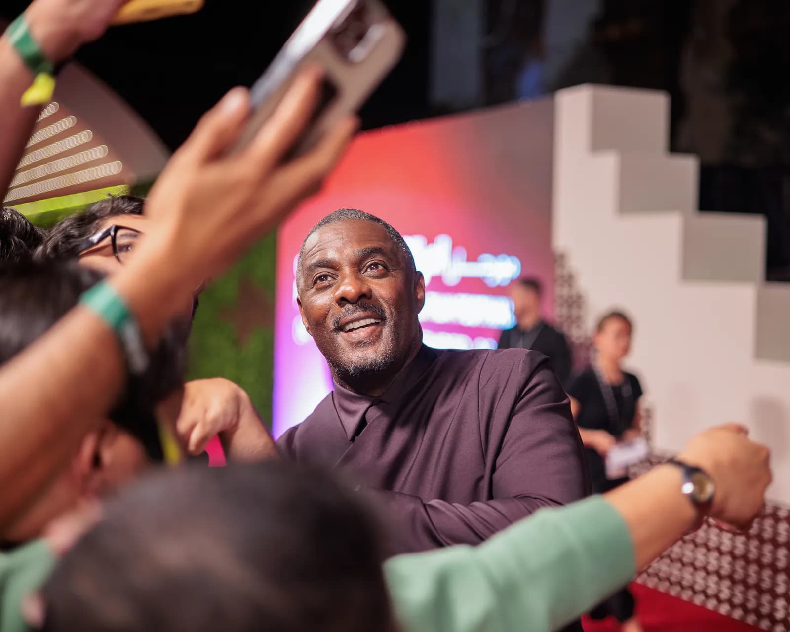 Idris Elba in a dark purple shirt smiles and looks up while surrounded by a blurred crowd with arms outstretched towards him and holding cell phones.