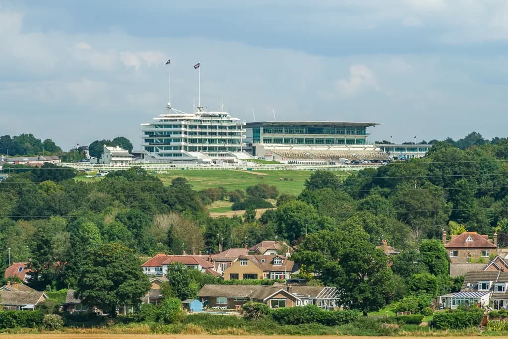 A large, modern white building with multiple tiers and green glass windows, along with a grandstand, sits atop a green hill overlooking a residential area with traditional houses and lush trees under a cloudy sky.