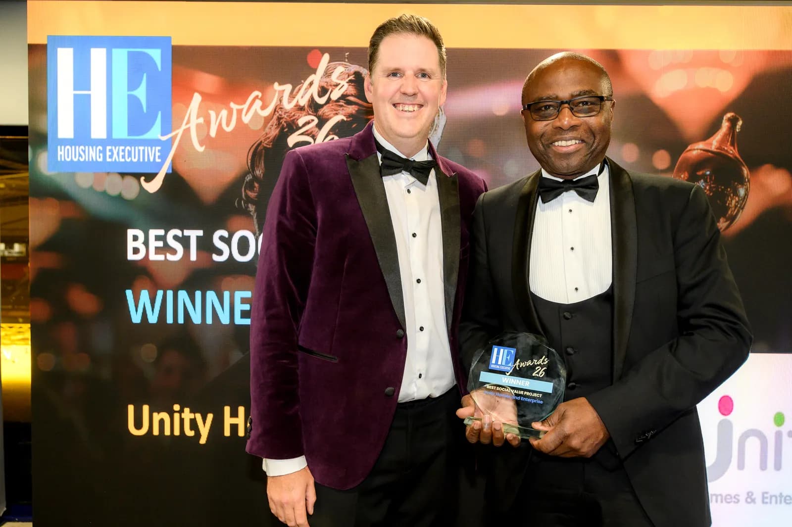 Two men in formal attire, one wearing a purple velvet jacket and the other a black suit, stand smiling while holding a clear award plaque that reads "HE Awards 26 Winner."