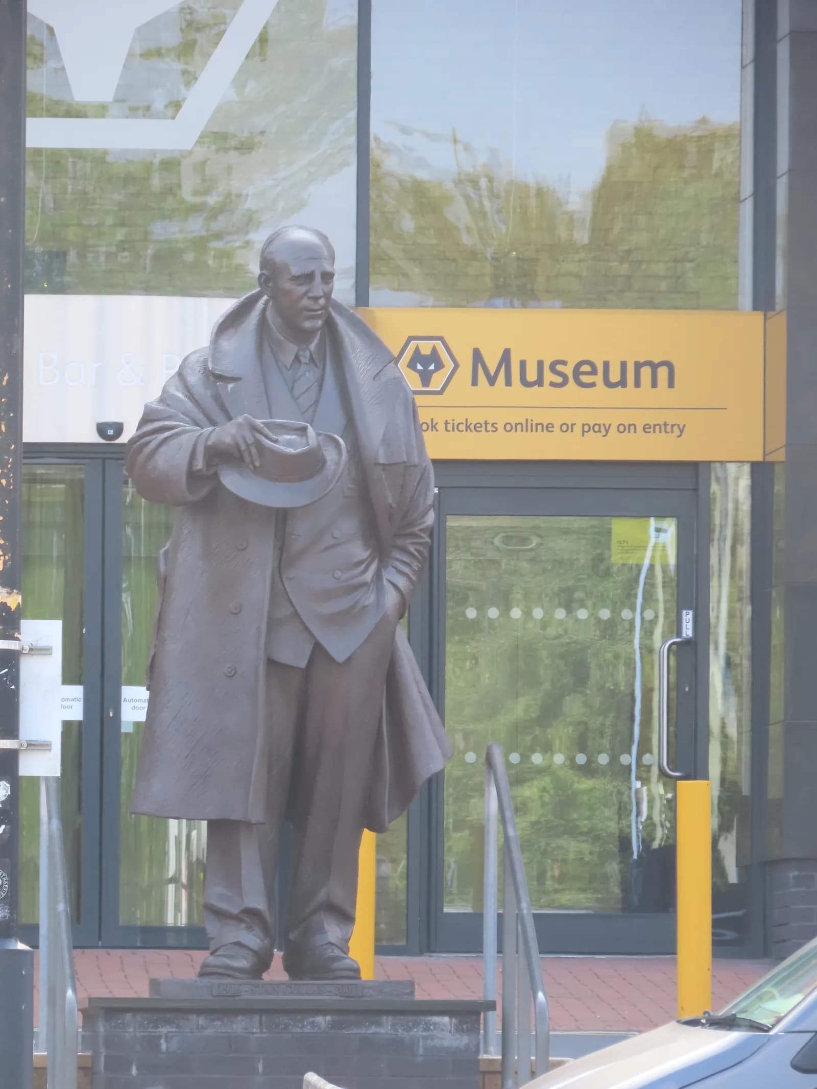 A bronze statue of a man in a trench coat and suit holding a fedora in his left hand, standing in front of a modern building with a yellow sign that reads "Museum" with a wolf head logo.