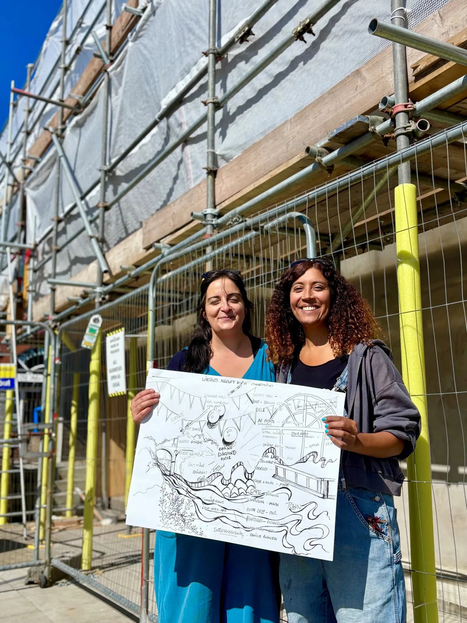 Two women, one with long dark hair and wearing a blue dress, the other with curly brown hair and wearing a black top and denim overalls, stand smiling and holding up a hand-drawn poster in front of scaffolding.