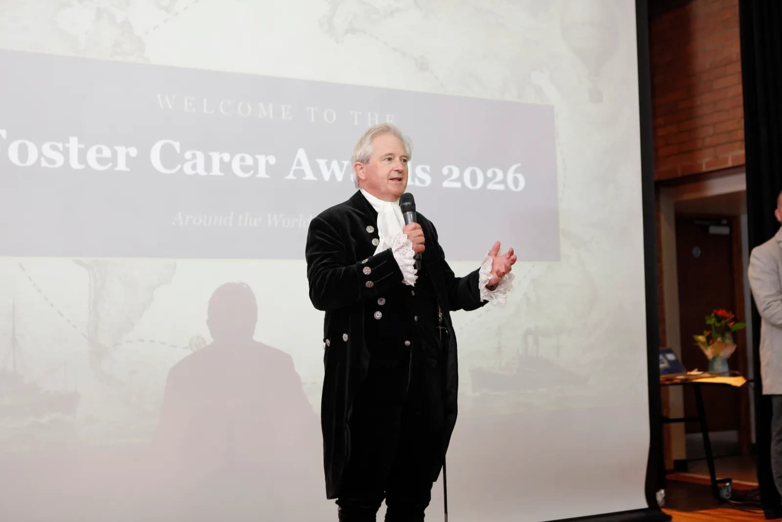 A man in historical attire, including a black long coat and ruffled white shirt, speaks into a microphone while gesturing with his left hand, standing in front of a screen displaying "Welcome to the Foster Carer Awards 2026."